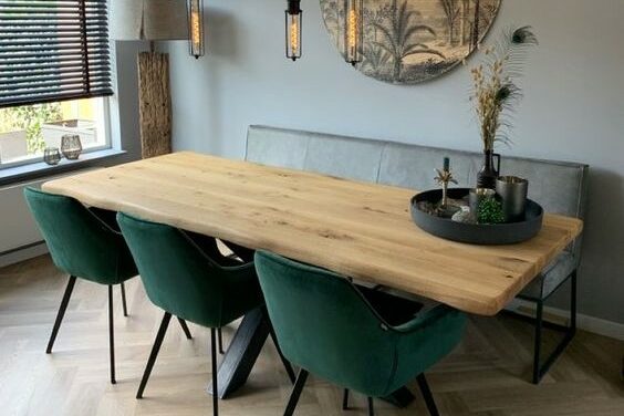 Salle à manger avec une table en bois rectangulaire, 3 fauteuils en velours vert bouteille et un banc en velours gris, sur un sol pointe de Hongrie, avec des luminaire suspendus noir et un tableau rond représentant des palmier. En décoration il y a un plateau rond noir avec des bougies et des plantes séchées.