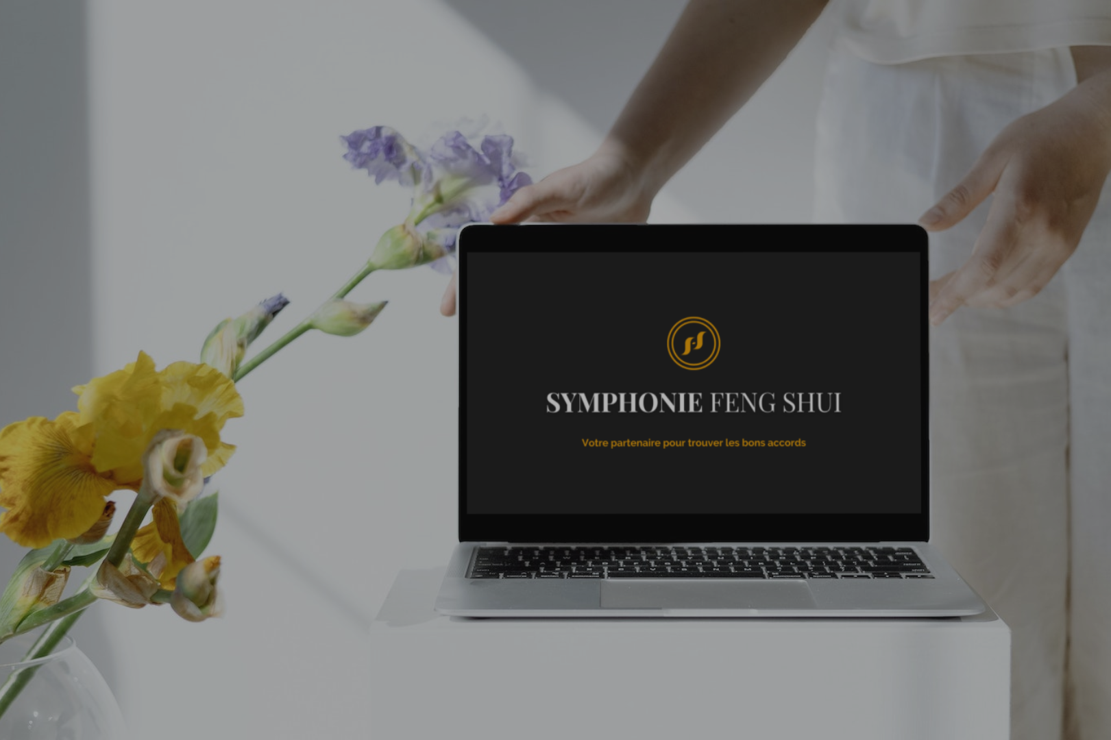 Ordinateur portable tenu par une femme avec le logo de Symphonie Feng-Shui, sur fond blanc et en présence de fleurs jaune et violette -Feng Shui Audit Aménagement intérieur Paris.